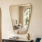 Handmade wavy top brass wall mirror with tapered sides and solid brass frame displayed above bedroom console.